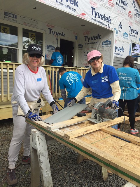  A group of Habitat for Humanity volunteers pose together on a construction site, wearing hard hats and smiling. Wooden framing and tools are visible behind them, highlighting active homebuilding efforts