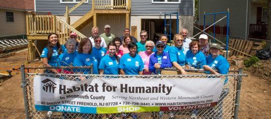 Habitat for Humanity volunteers stand smiling in front of a newly built home, holding a framed certificate. They are wearing branded T-shirts and hard hats, with construction tools visible in the background.